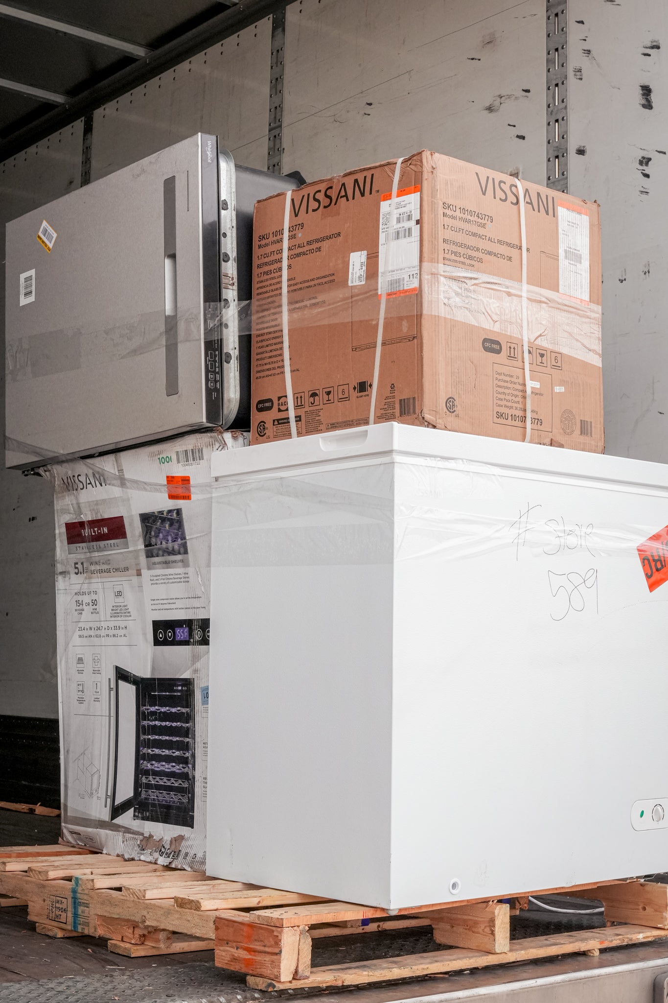 Stack of stacked appliances including a microwave and refrigerator on a pallet with visible brand names.